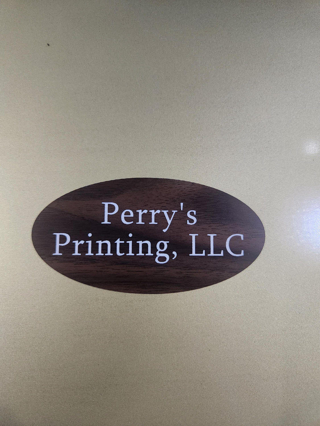 Wood-style engraved oval sign with white text reading 'Perry's Printing, LLC' on a gold background.