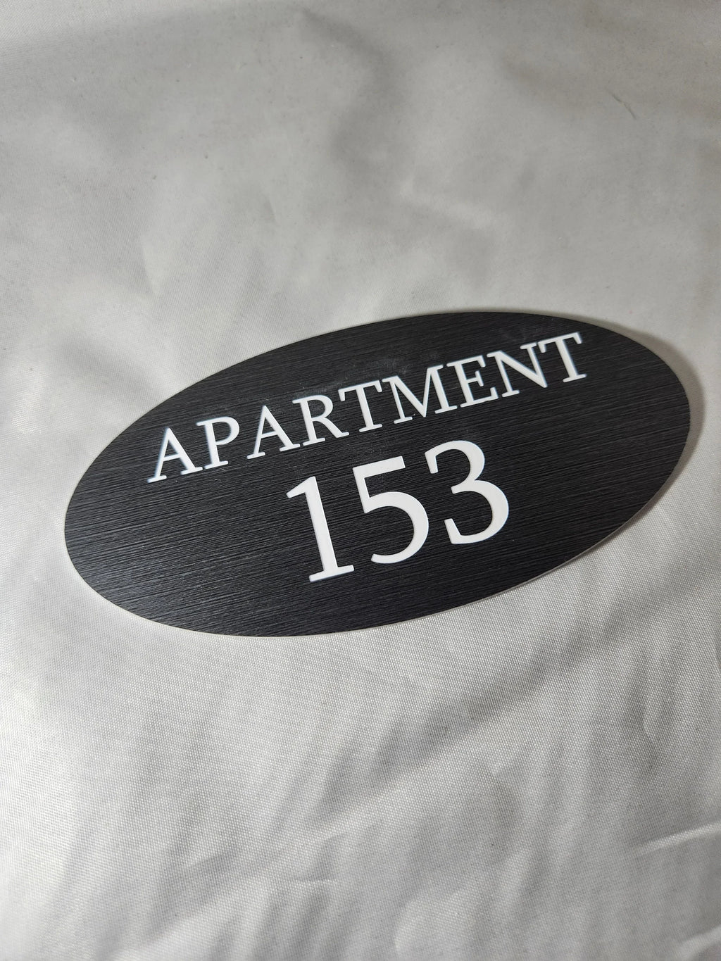 Oval black apartment number sign with white text reading APARTMENT 153 on fabric background