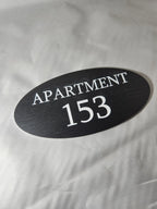 Oval black apartment number sign with white text reading APARTMENT 153 on fabric background