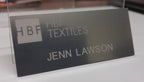 Laser engraved metallic nameplate for HBF Textiles with Jenn Lawson's name, on white surface.