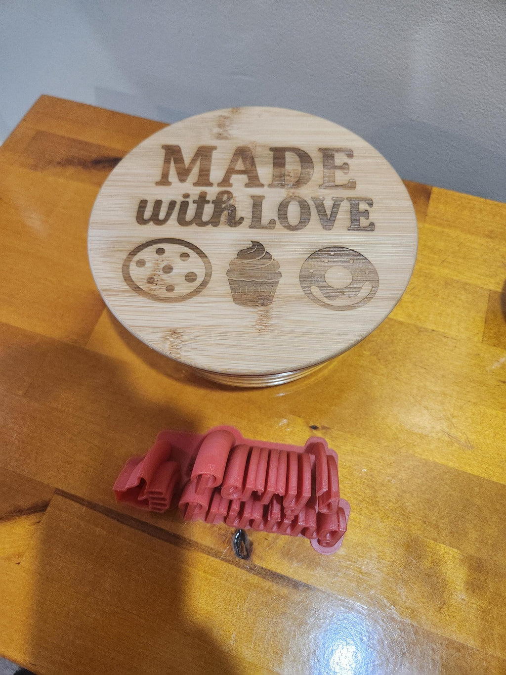 Laser-engraved bamboo lid with 'Made with Love' and cookie, cupcake, donut icons, next to 3D printed red text mold on wooden table.