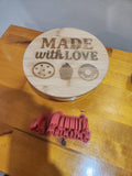 Laser-engraved bamboo lid with 'Made with Love' and cookie, cupcake, donut icons, next to 3D printed red text mold on wooden table.