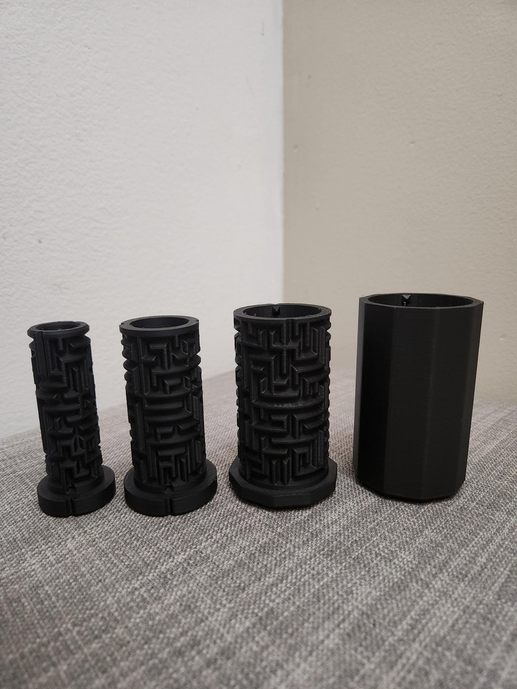 Four black 3D printed cylindrical puzzle boxes with maze patterns on a gray fabric surface