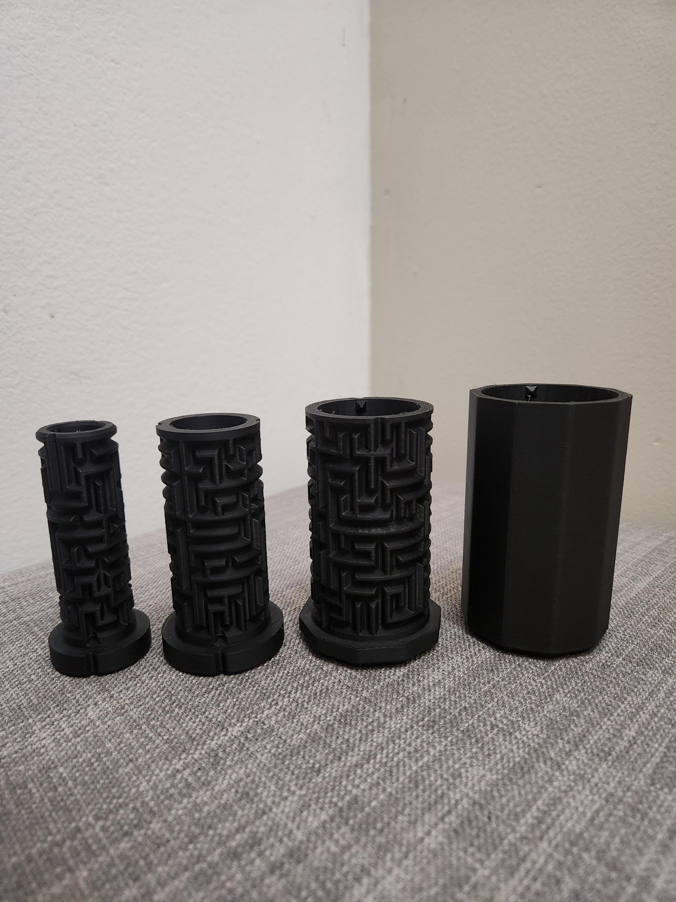 Four black 3D printed cylindrical puzzle boxes with maze patterns on a gray fabric surface