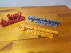 3D printed Instagram and business name signs in red, silver, and yellow on a wooden table.