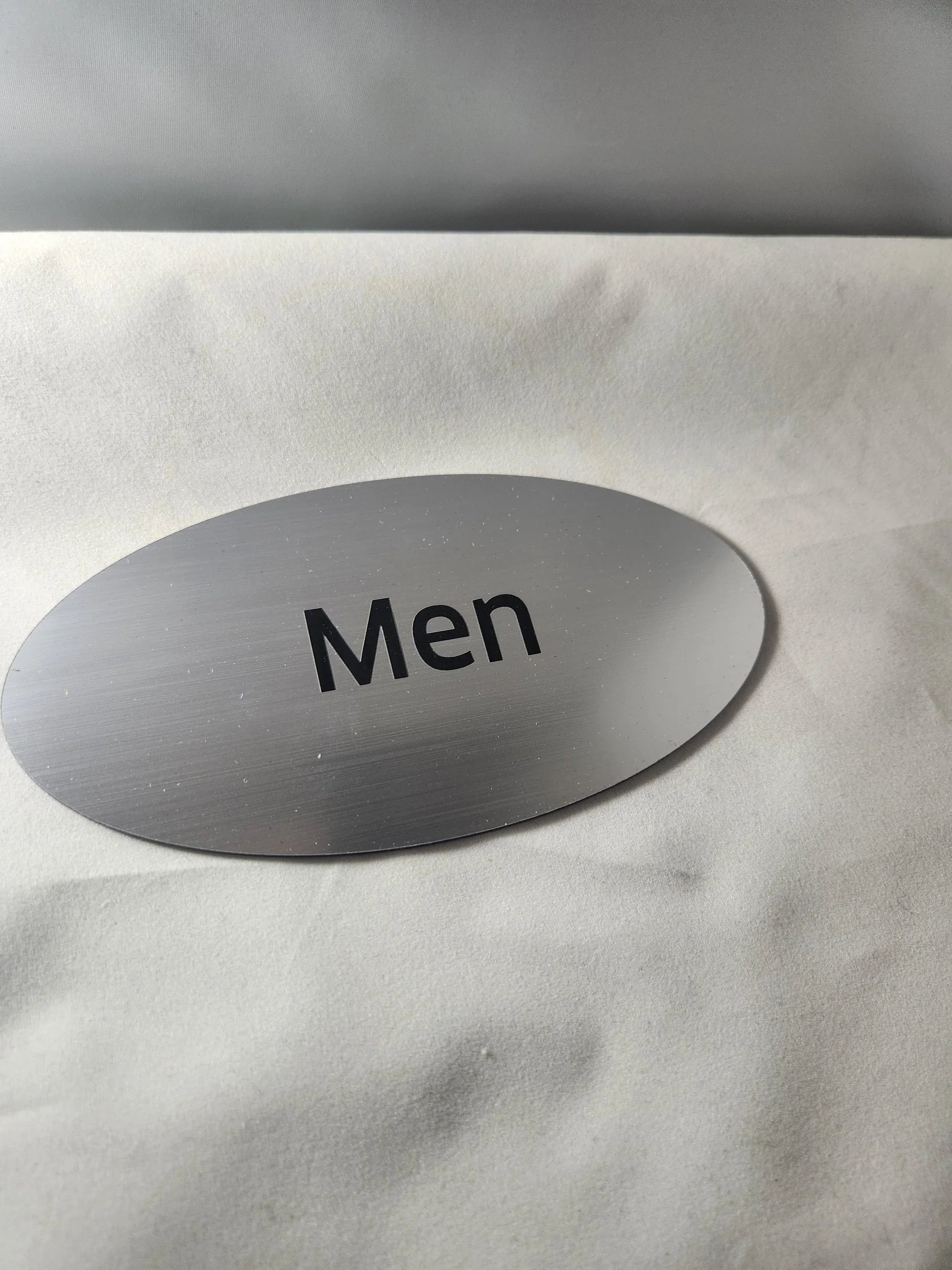 Brushed aluminum oval men’s restroom sign with black lettering on a white background