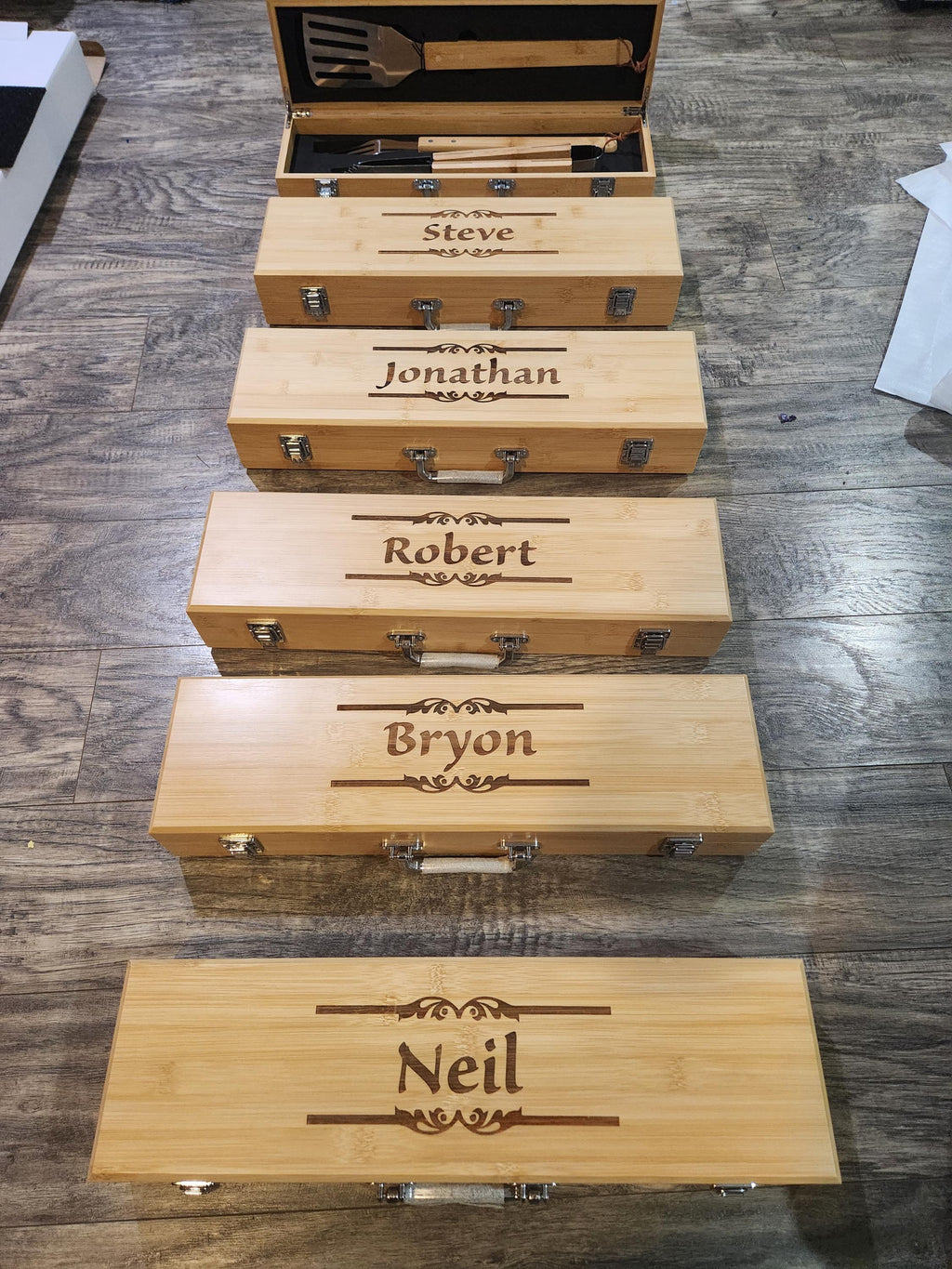 Custom engraved bamboo BBQ tool boxes with personalized names, arranged on wood floor