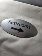 Oval brushed metal restroom sign with arrow, laser engraved, on light fabric background