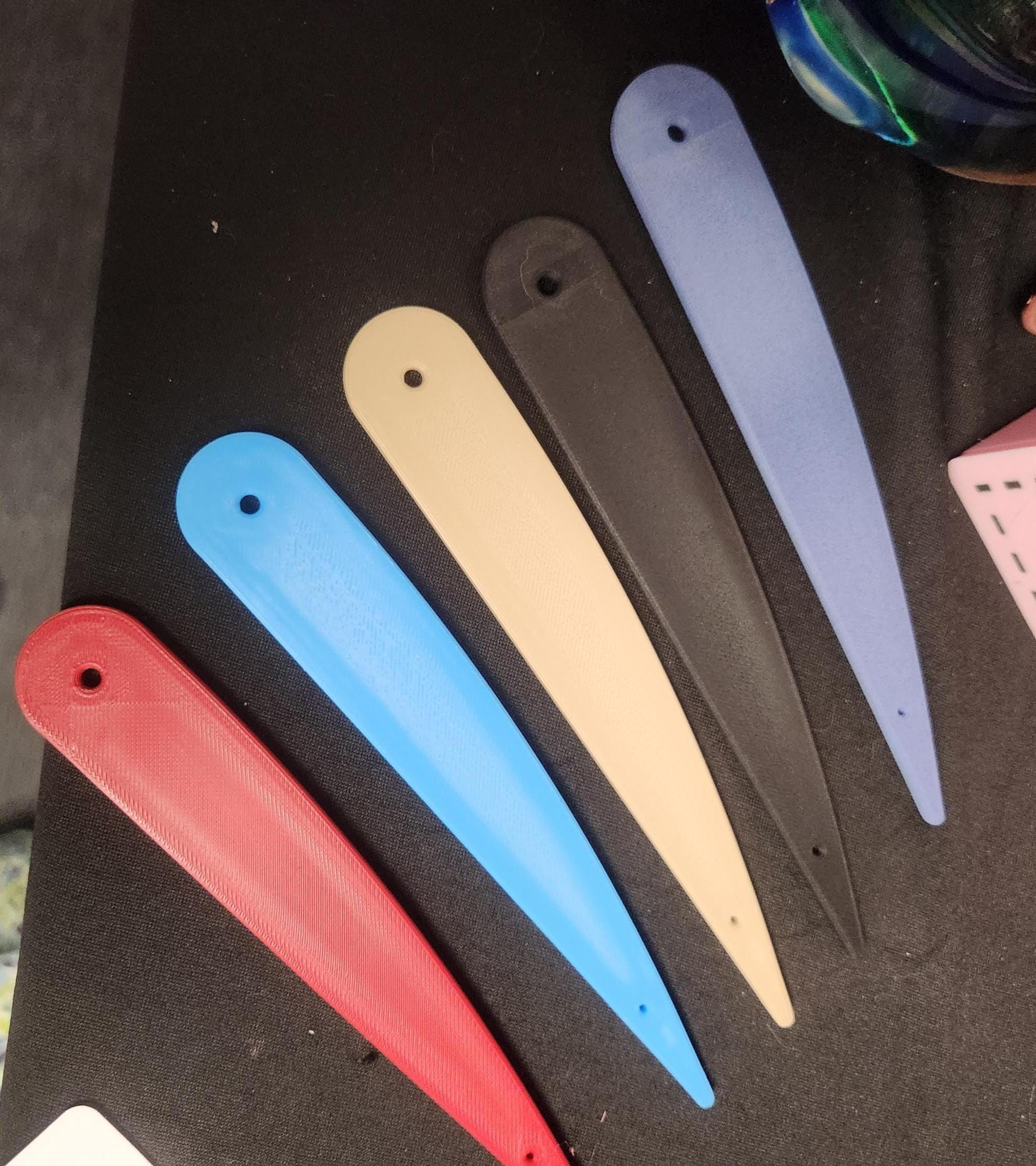 Assorted 3D printed plastic letter openers in red, blue, beige, black, and lavender on a black surface.
