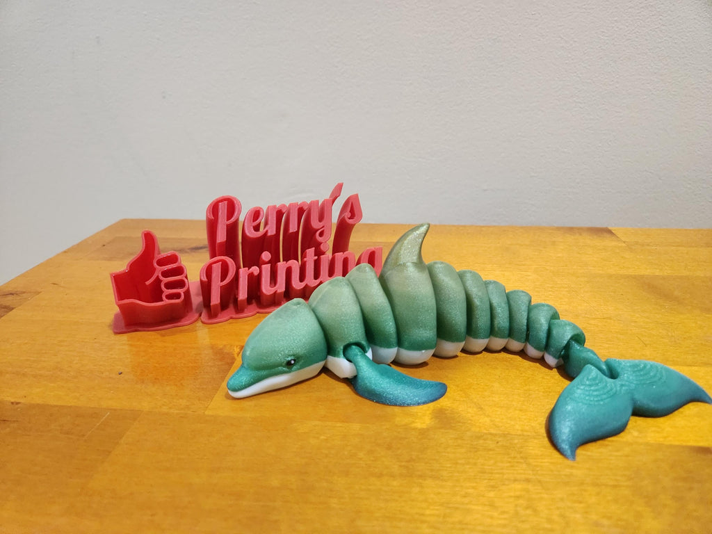 3D printed flexible dolphin figure and red Perry's Printing sign with thumbs up on wood table