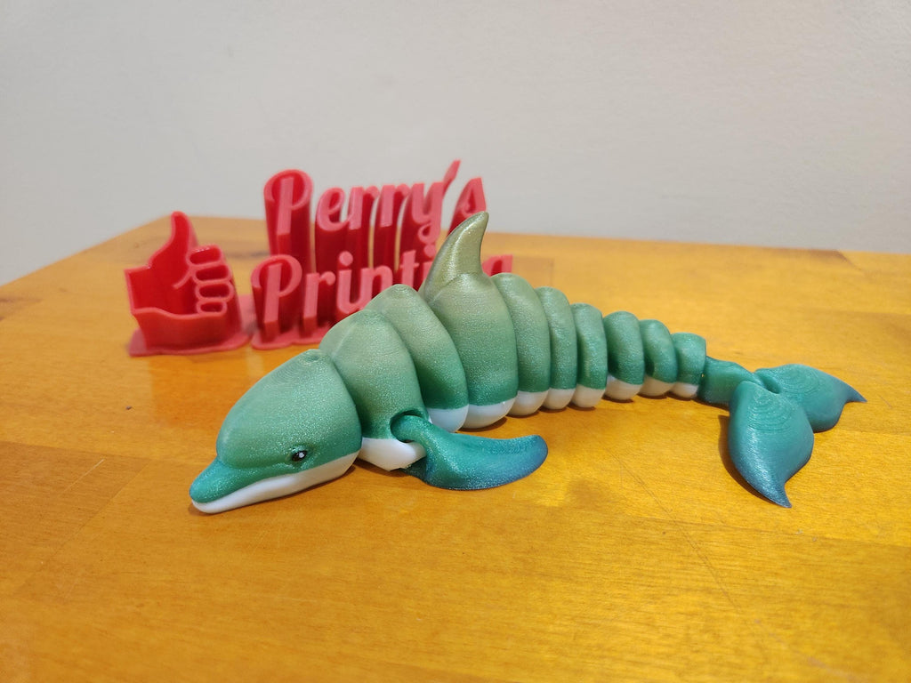 3D printed articulated dolphin figurine on wood surface, red printed thumbs up sign in background