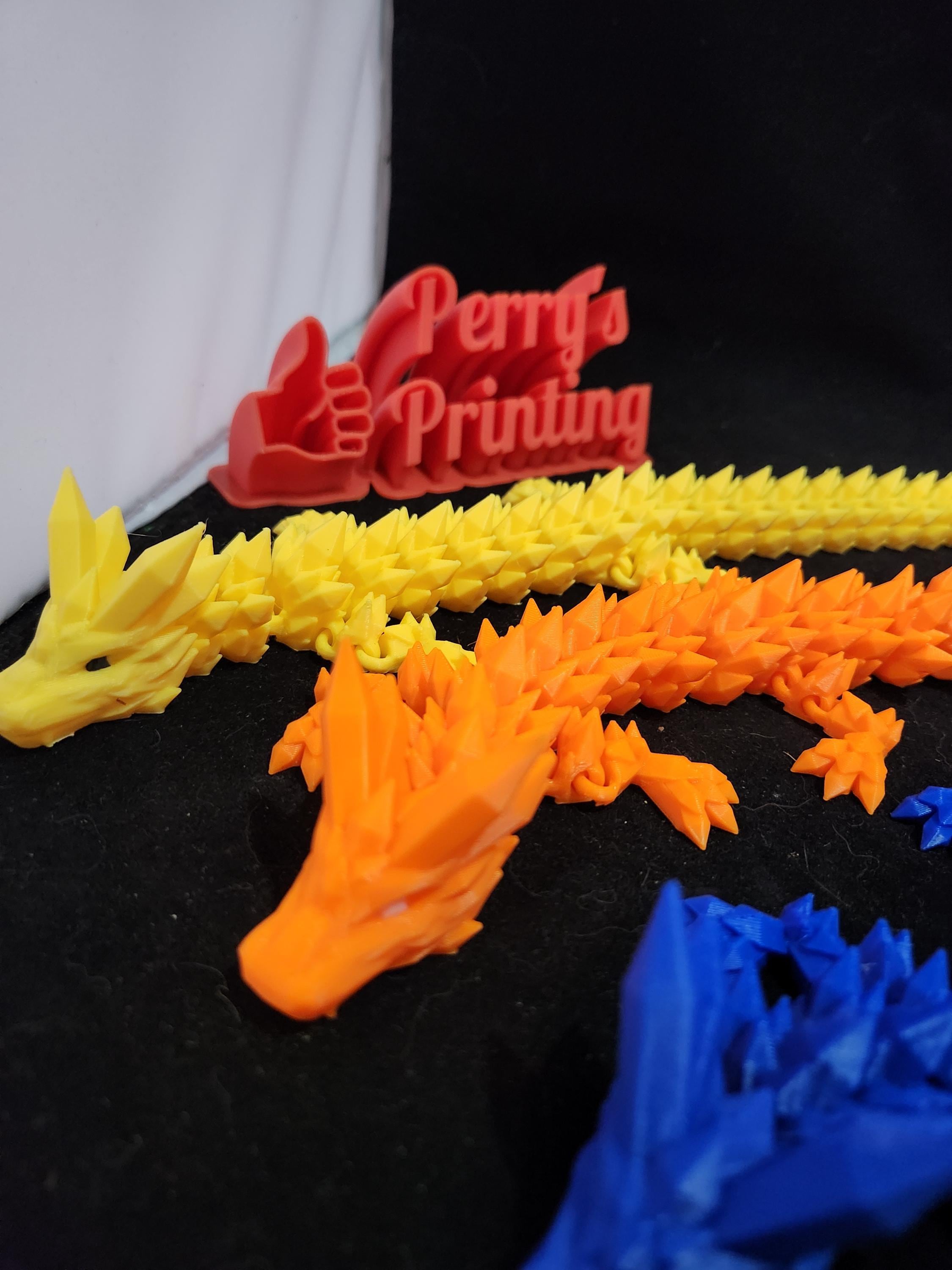 Colorful 3D printed dragon figurines on black surface with Perry's Printing sign in background