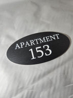 Oval black apartment door number sign with 'APARTMENT 153' in white text, custom signage