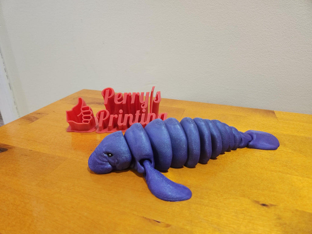 Purple 3D printed articulated manatee on wood surface, red 3D printed sign in background