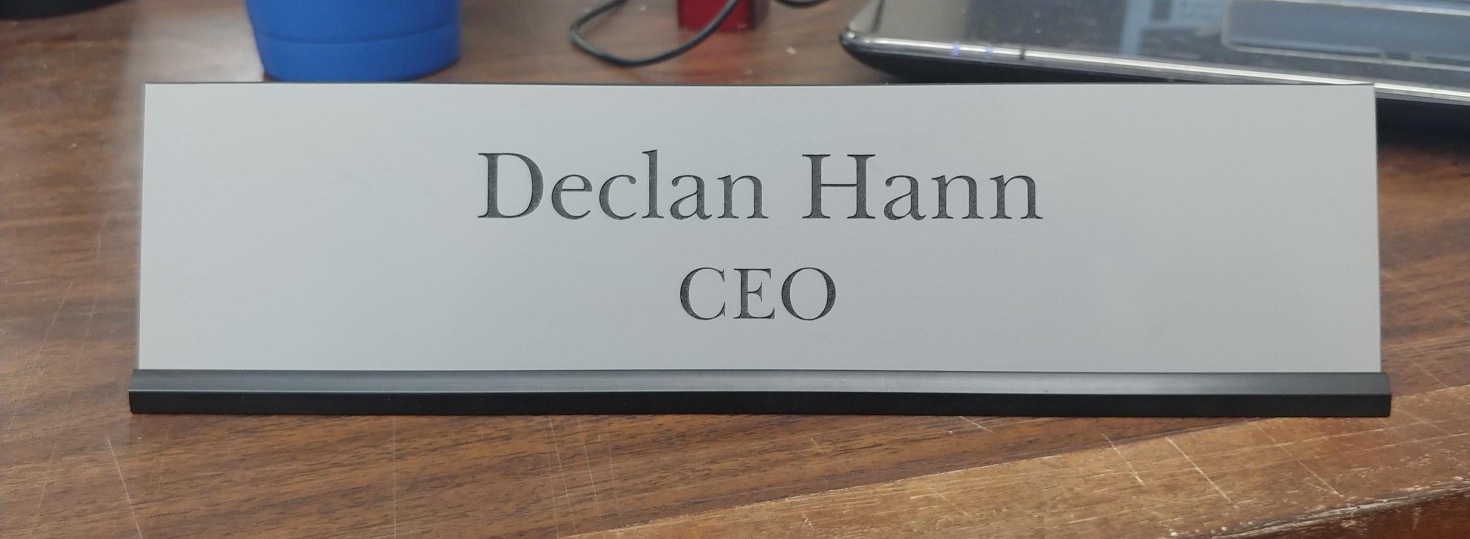 Custom desk nameplate with black text CEO title, silver background, black holder, on wood desk