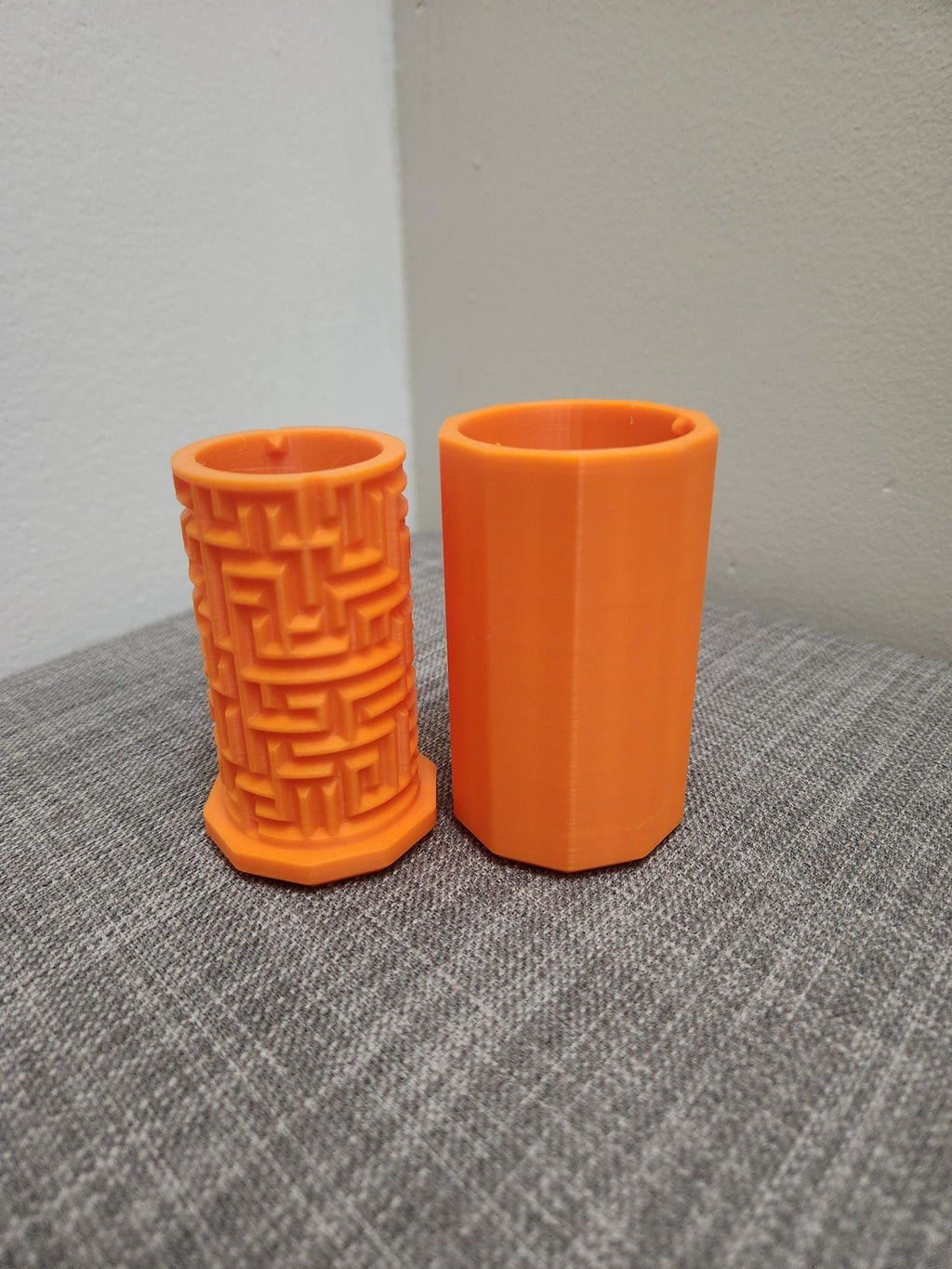 Two orange 3D printed pen holders, one with a geometric maze pattern, on a gray fabric surface