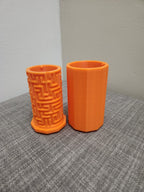 Two orange 3D printed pen holders, one with a geometric maze pattern, on a gray fabric surface