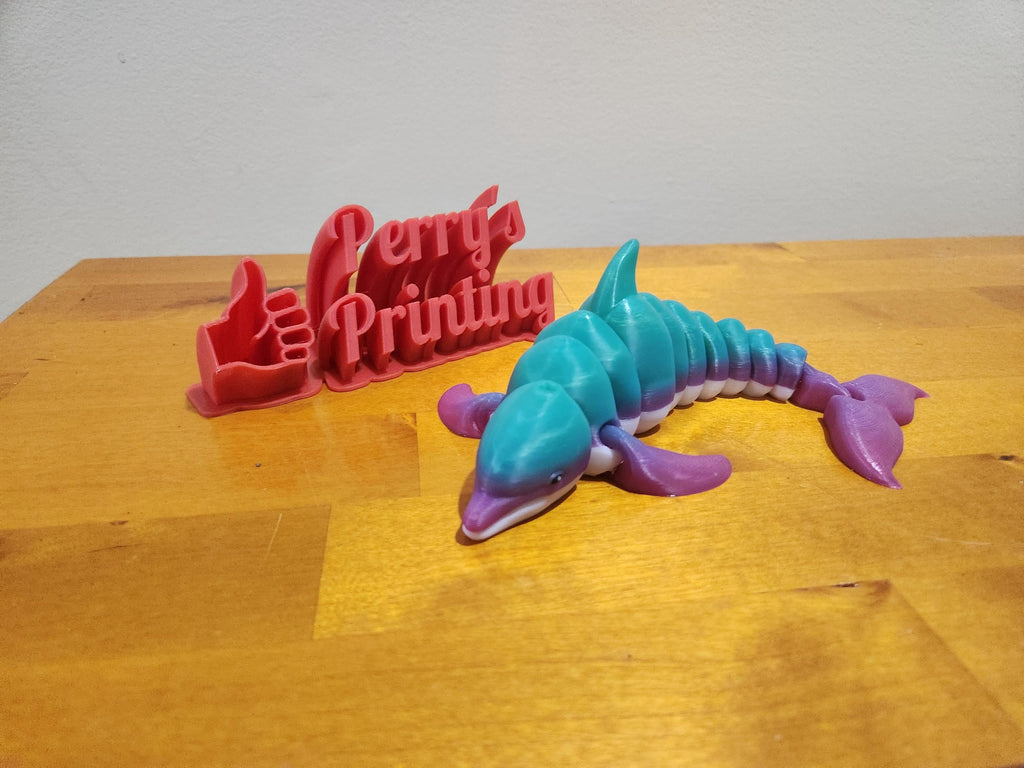 3D printed flexible whale toy in blue and purple next to red Perry's Printing sign on wood table