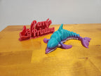 3D printed flexible whale toy in blue and purple next to red Perry's Printing sign on wood table