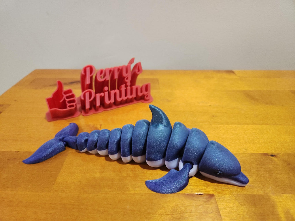 3D printed jointed blue dolphin figurine on wooden surface with red Perry's Printing sign