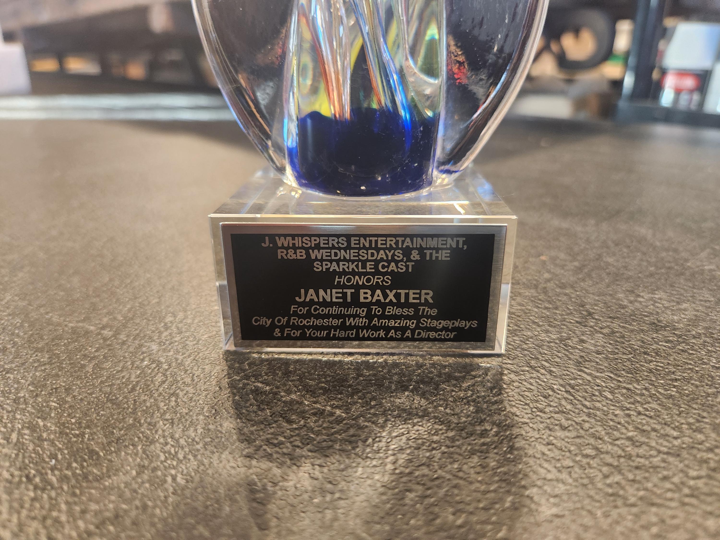 Custom engraved award with blue glass detail on a textured tabletop, honoring Janet Baxter.