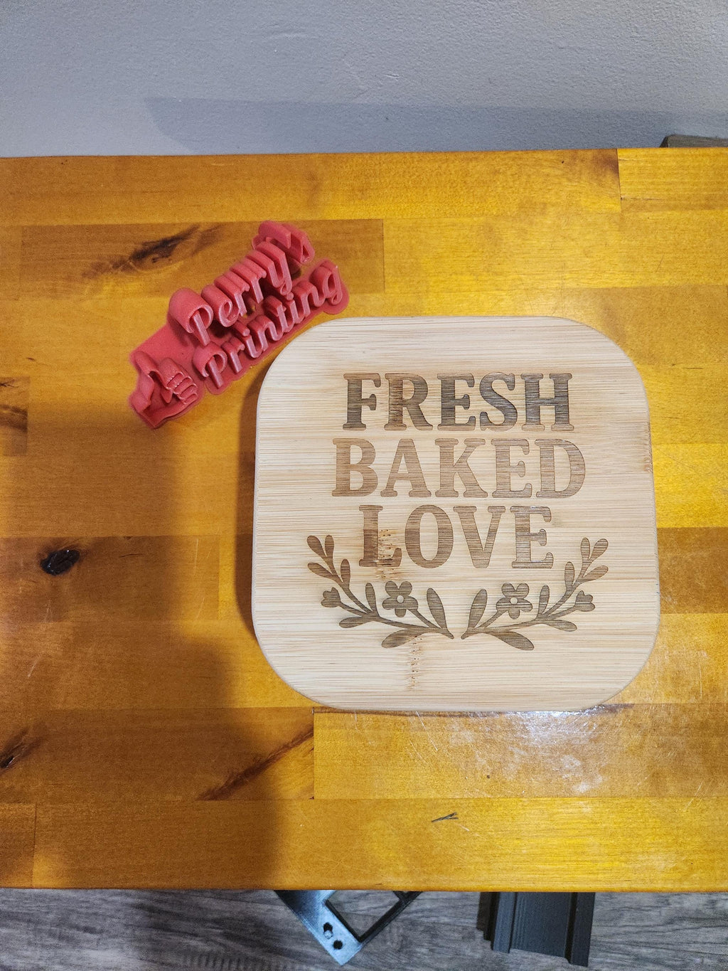 Laser engraved bamboo sign with 'Fresh Baked Love' and a red 3D printed sign on wooden table.