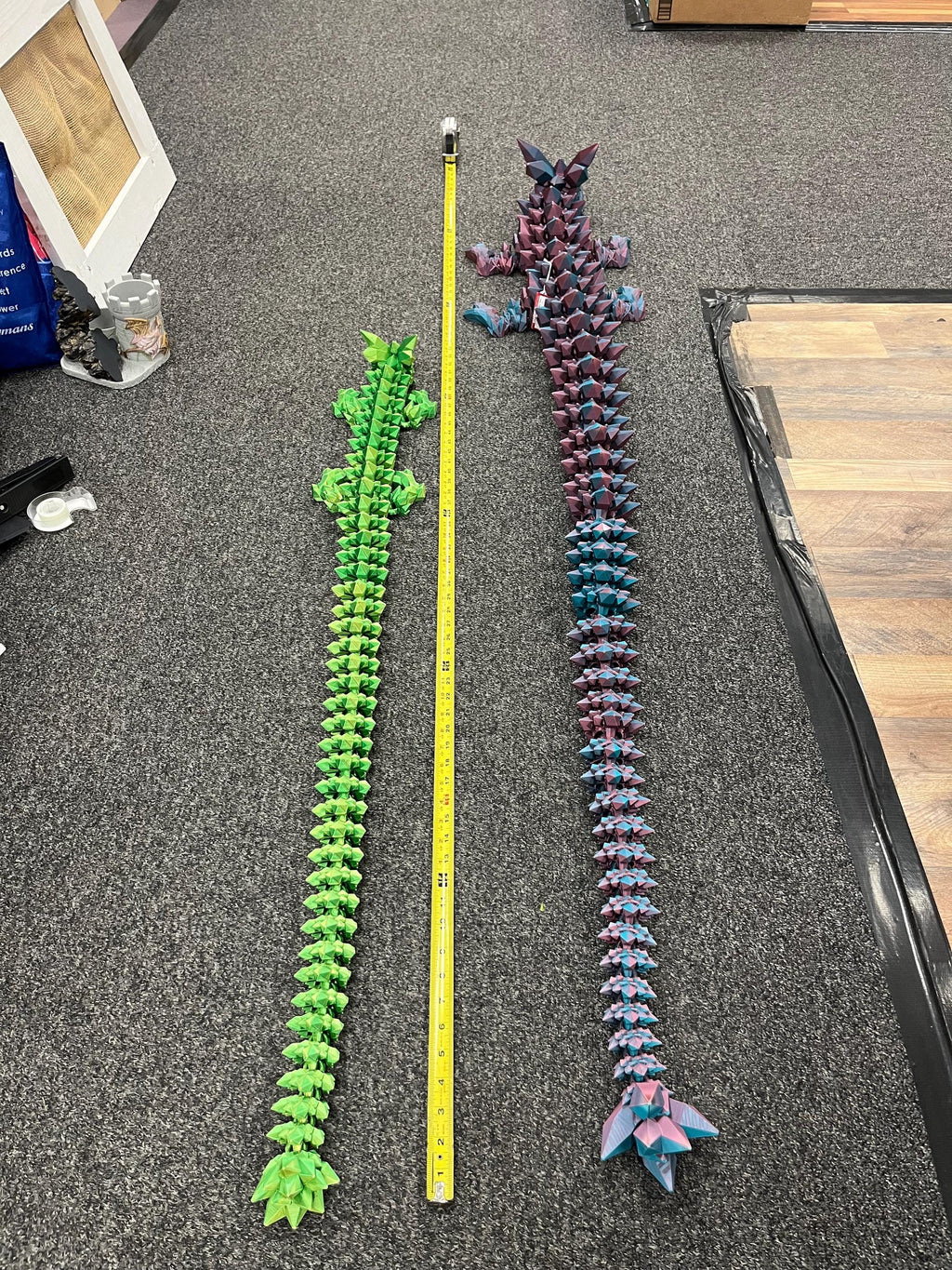 Two large 3D printed articulated dragon figures, one green and one purple-blue, beside a tape measure on a carpeted floor.
