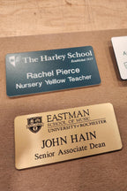 Custom engraved name badges on a table, including gold and blue school staff tags.