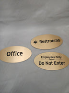 Gold oval engraved signs reading Restrooms, Office, and Employees Only Do Not Enter on gray fabric